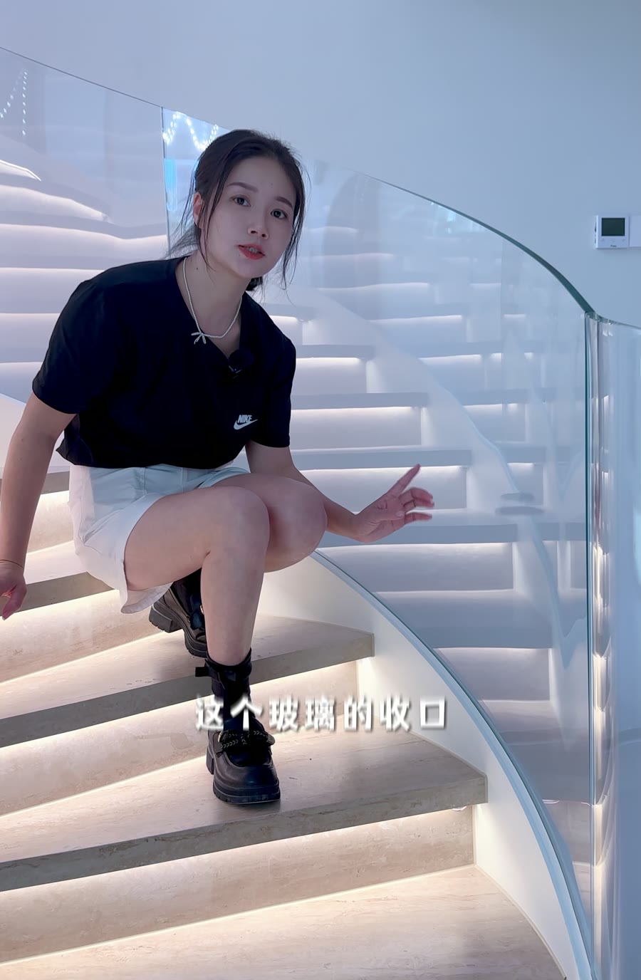 女性蹲在弧形楼梯踏步上，手指向玻璃栏板与踏步交接的收口位置，旁边是透明玻璃栏板。